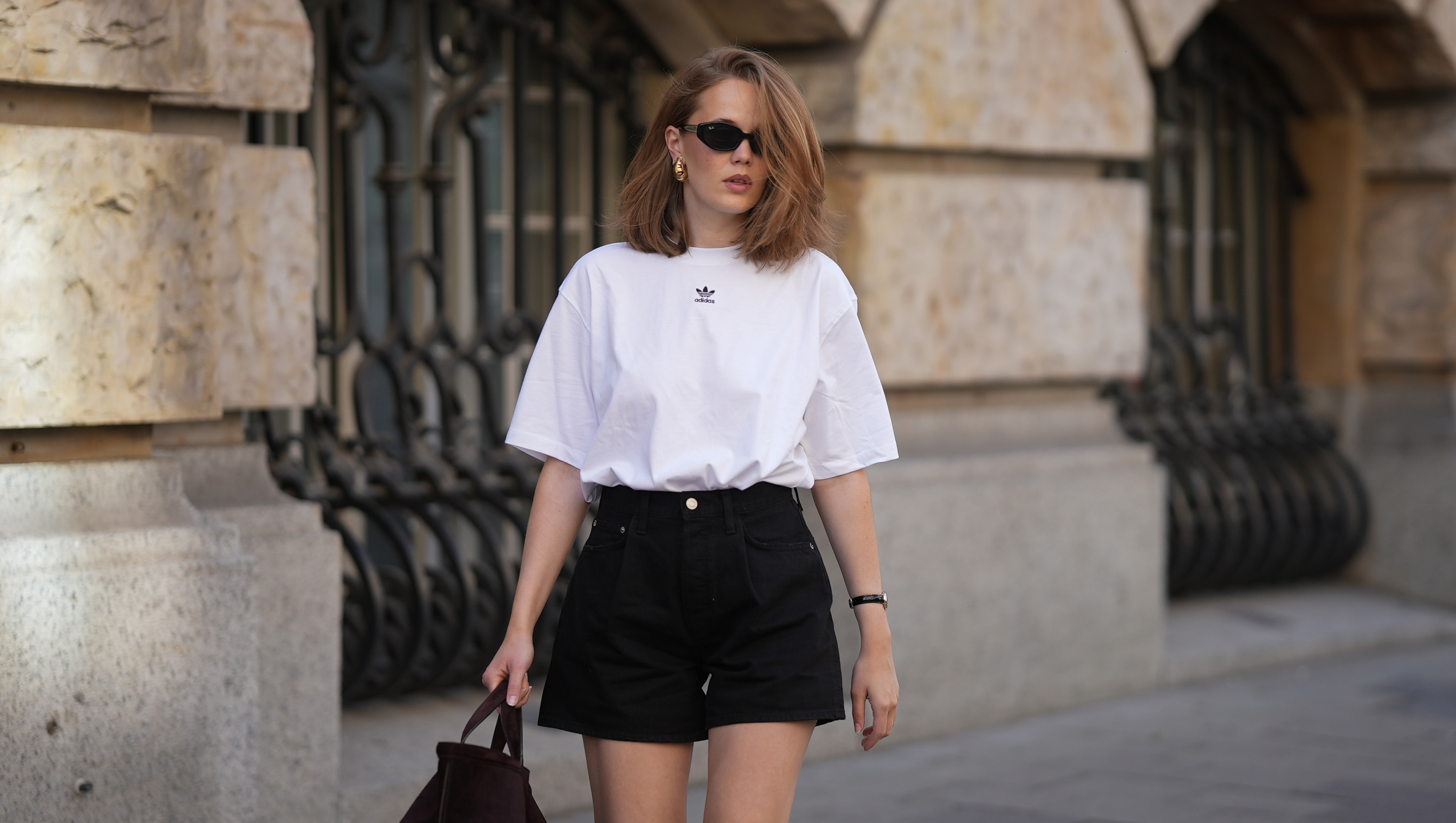 MUNICH, GERMANY - JUNE 8: Pia Riegel is seen wearing an oversized white t-shirt by Adidas; black high-waisted denim shorts by Agolde; a dark brown suede wrist bag with a soft slouchy shape and short rounded handles by COS; black oval sunglasses by Ray-Ban; silver headphones by Apple; a gold-tone wristwatch with a light dial and black leather strap; gold hoop earrings; shoulder-length softly tousled hair; with natural make up on June 8, 2026 in Munich, Germany. (Photo by Moritz Scholz/Getty Images)