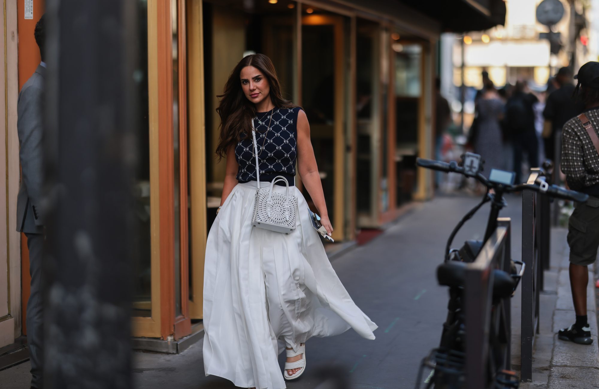 PARIS, FRANCE - JULY 05: A guest seen wearing gold pendant necklaces, a black bow pattern knit sweater vest, a white oversized pleated long skirt/pants, a white leather hole pattern crossbody bag and white leather sandals, outside the Giorgio Armani Prive show, during Paris Fashion Week - Haute Couture Fall Winter 2022 2023, on July 05, 2022 in Paris, France. (Photo by Jeremy Moeller/Getty Images)