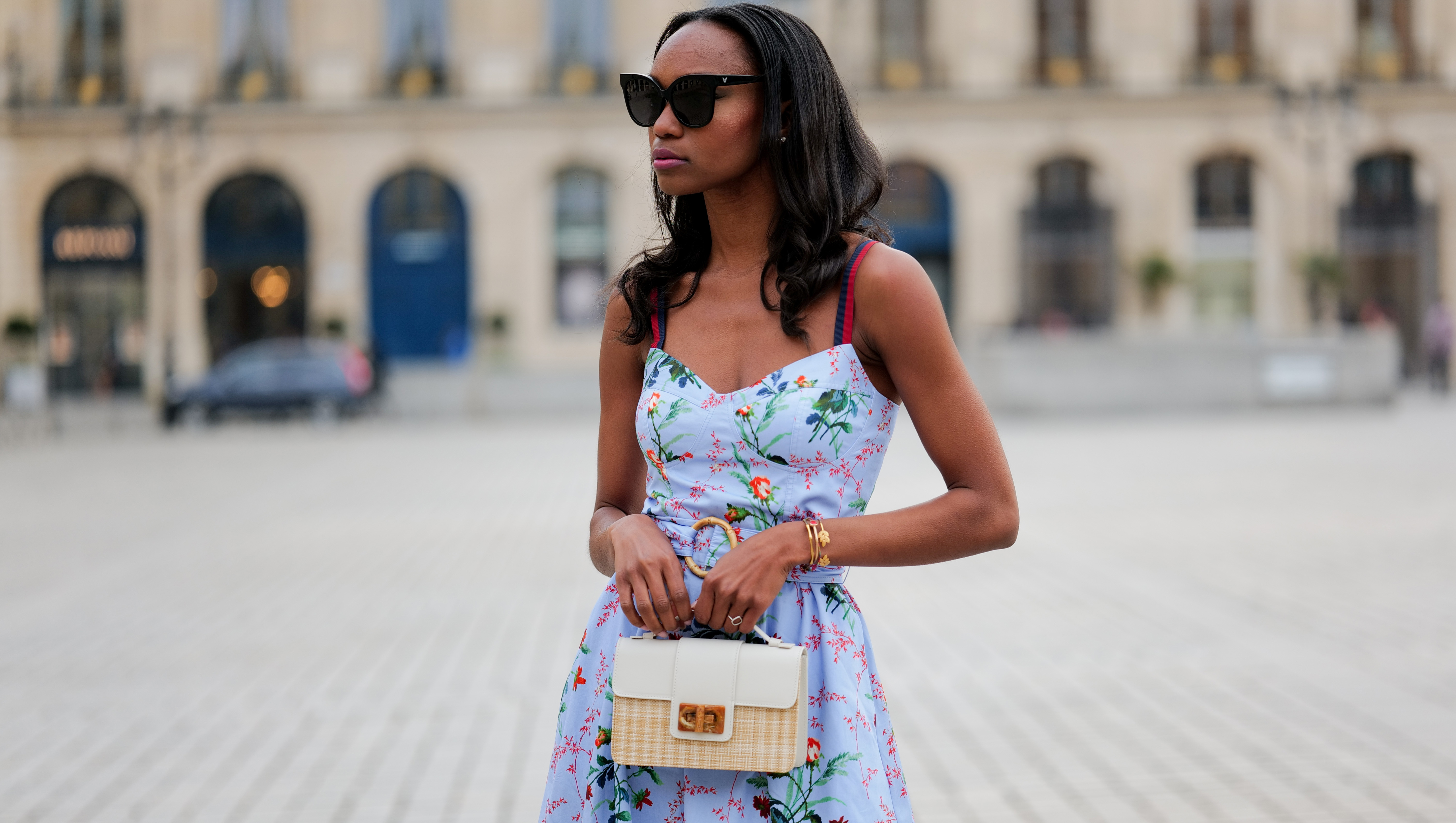 PARIS, FRANCE - APRIL 29: Emilie Joseph wears black large sunglasses, crystal earrings, a pale blue with red / green / navy blue / beige flower print pattern heart neck / tank-top / belted / midi dress, a beige wickers and white shiny leather bi-material handbag, during a street style fashion photo session, on April 29, 2023 in Paris, France. (Photo by Edward Berthelot/Getty Images)