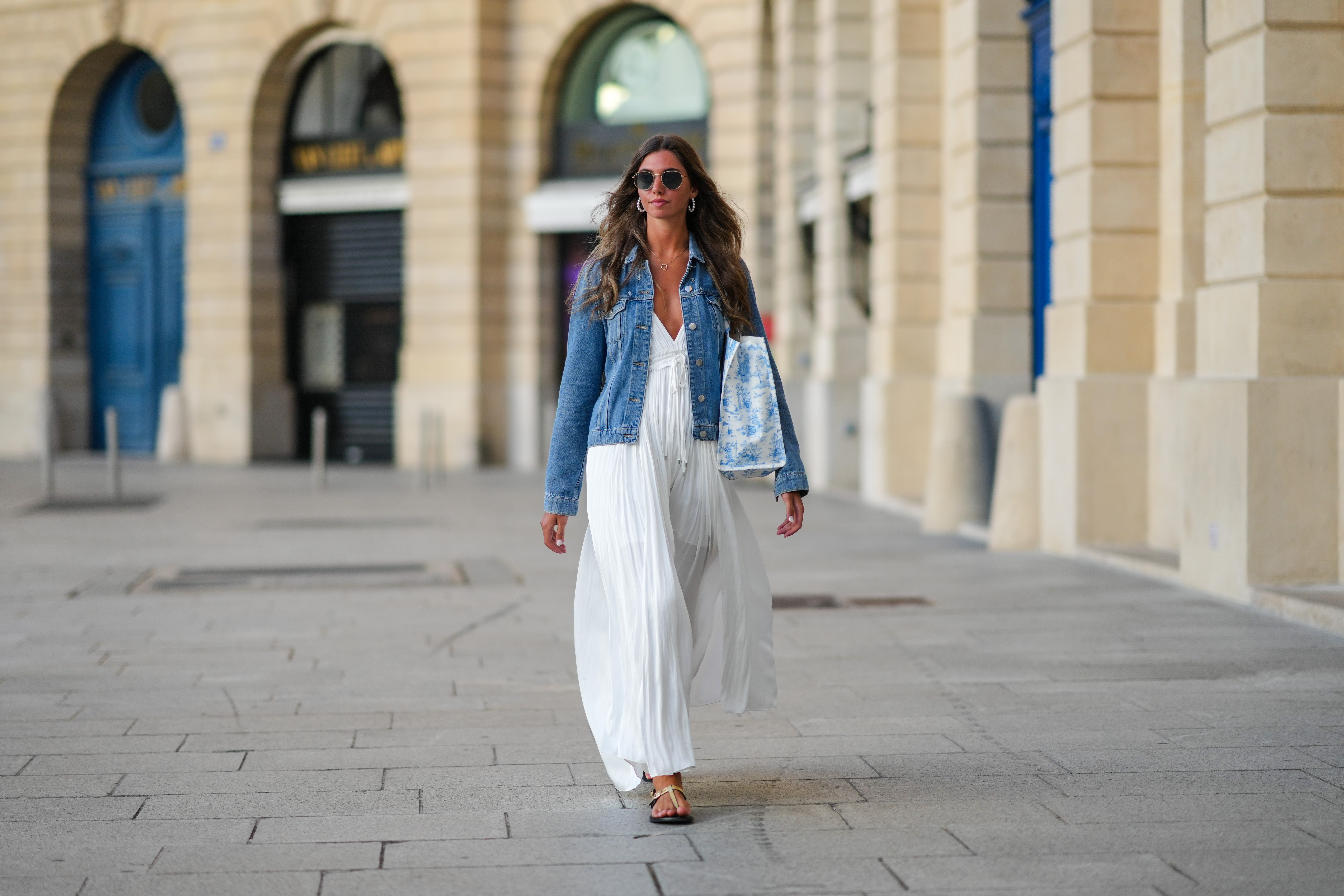 Amanda Derhy wears earrings, sunglasses, a blue denim jacket, a white summer maxi pleated low-neck slit off-shoulder dress, sandals, during a street style fashion photo session, on August 20, 2022 in Paris, France.