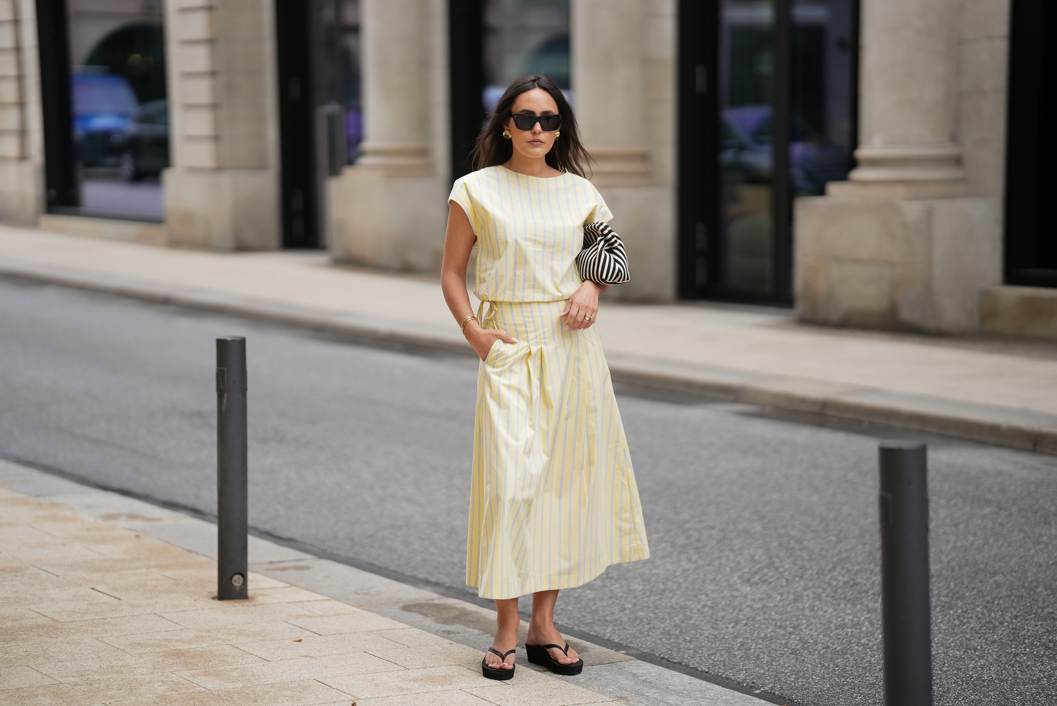 Laura Noltemeyer is seen wearing a yellow striped two-piece set with a sleeveless draped top and matching midi skirt by Sézane; black flip-flop sandals by Amazon; a black-and-white striped Porter clutch bag; black cat-eye sunglasses by Saint Laurent; nude-colored fingernails and toenails; gold jewelry by H&M and COS consisting of earrings, bangles and rings; natural makeup; her dark brown hair is worn in soft waves with a middle part on May 5, 2026 in Hamburg, Germany.