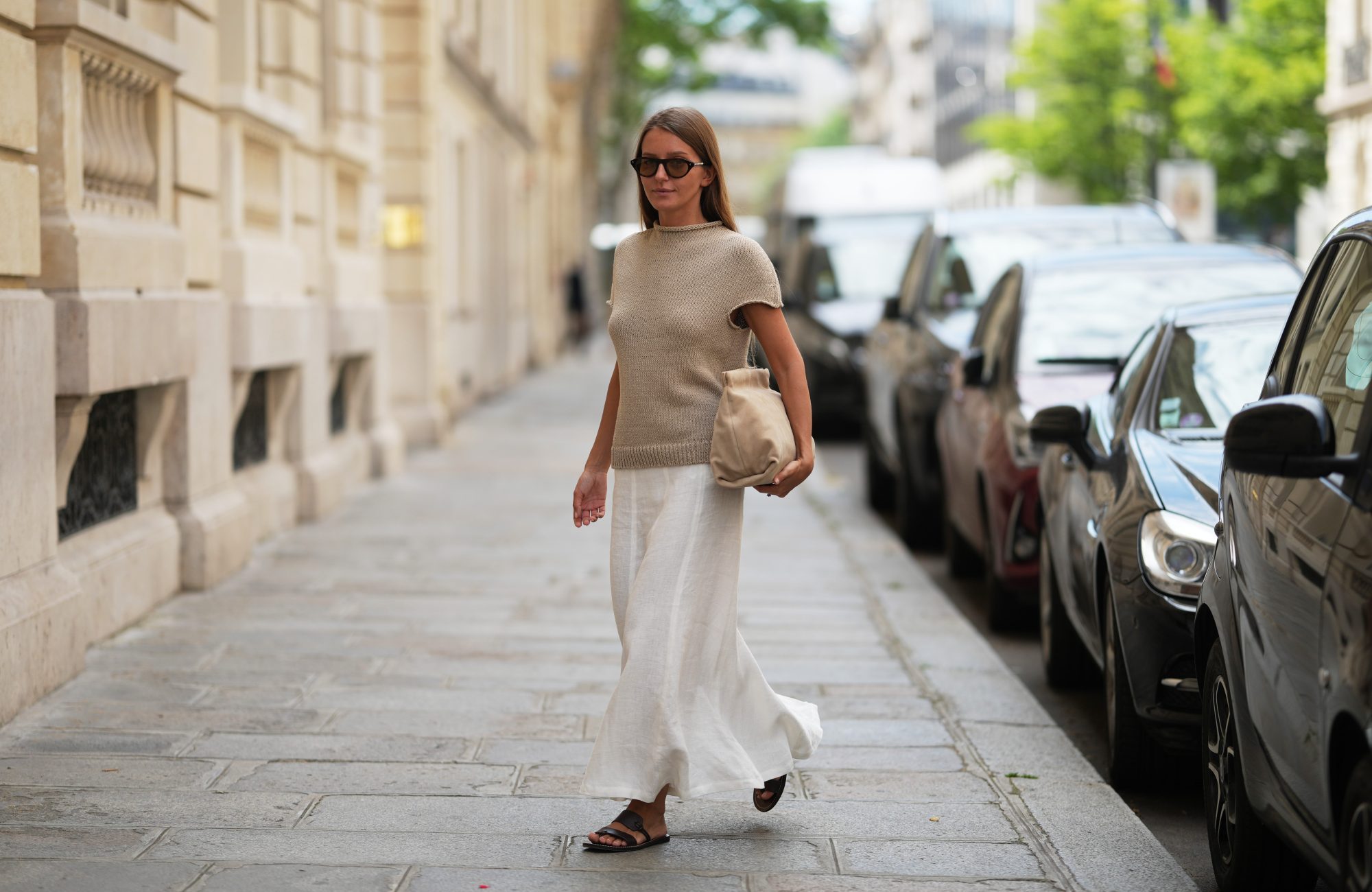 Diane Batoukina wears long straight brown hair, black rectangular sunglasses by Montblanc, a beige chunky knit short-sleeve top by Massimo Dutti, a white maxi skirt by Massimo Dutti, a beige clutch bag by Flattered, dark brown sandals leather shoes by Soeur, during a street style fashion photo session, on June 16, 2026 in Paris, France.
