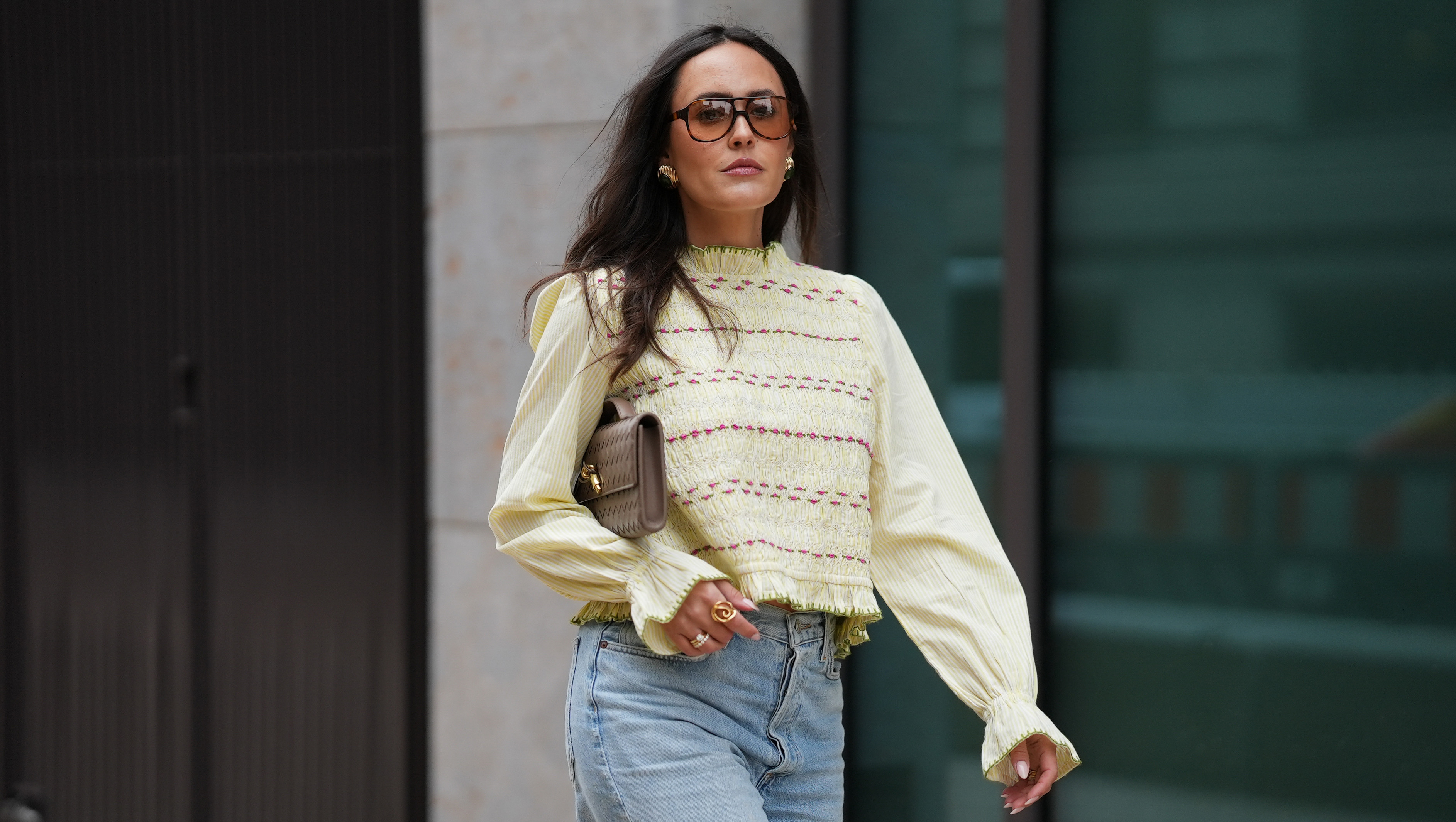 HAMBURG, GERMANY - MAY 5:Laura Noltemeyer is seen wearing the "Adame" blouse in a light lemon yellow a cotton fabric with a smocked bodice, pink and green embroidery, long balloon sleeves with ruffled cuffs and a back tie-bow closure from Sézane; light blue wide-leg denim jeans from Agolde; the "Andiamo" intrecciato woven leather clutch in pinecone with a top handle and gold hardware from Bottega Veneta; gold rings including a diamond ring; gold statement earrings with resin stone detail from Mango; oversized aviator sunglasses with a tortoiseshell patterned frame from Corlin; her dark brown hair is worn loose and wavy on May 5, 2026 in Hamburg, Germany. (Photo by Moritz Scholz/Getty Images)