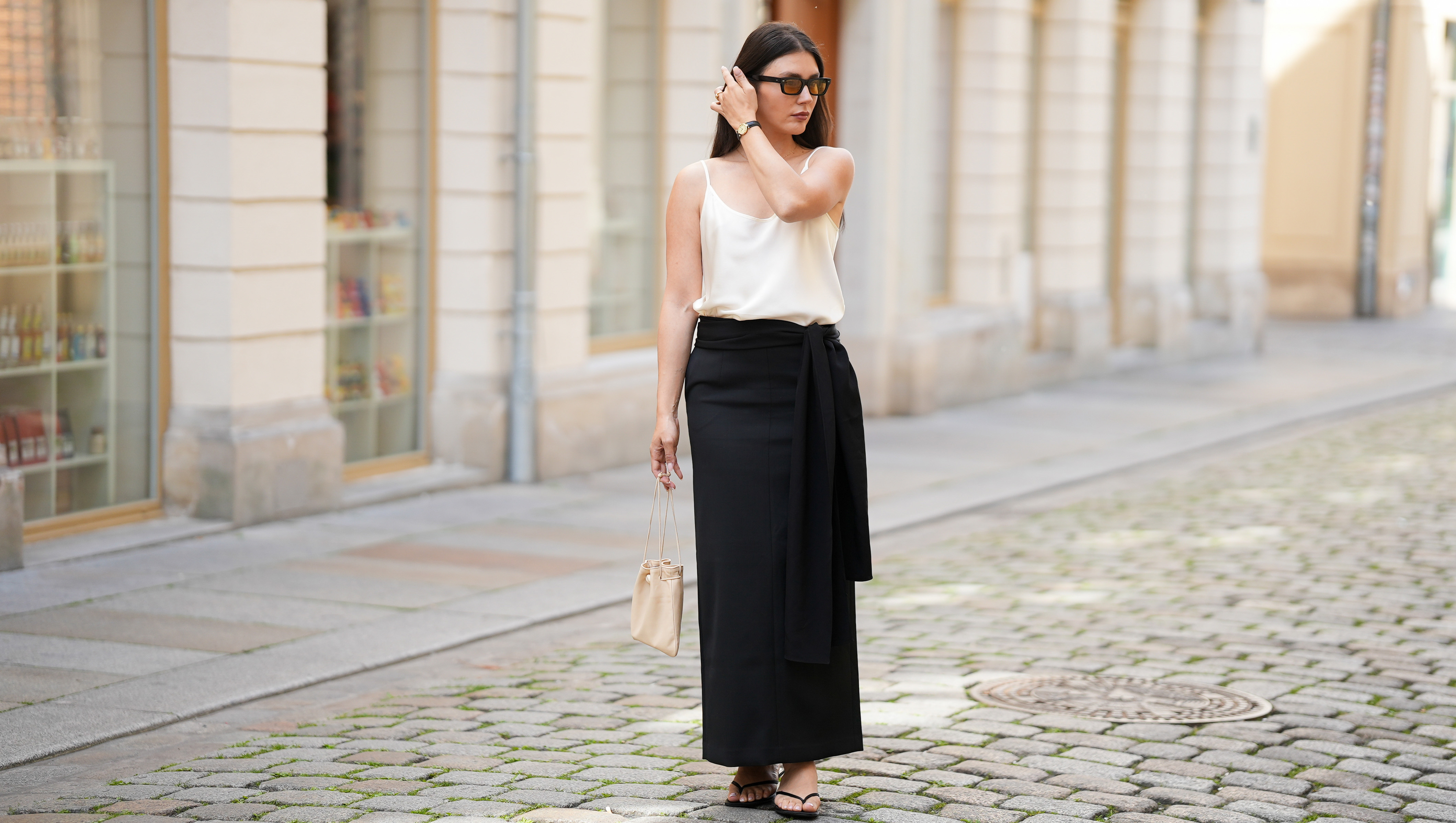 BERLIN, GERMANY - MAY 24: Milena Karl is seen wearing a white satin camisole top with spaghetti straps and a relaxed silhouette from Yael; a black satin maxi skirt with a high waist, straight silhouette and asymmetric wrap panel detail from Almada; a black lightweight scarf draped at the waist from Almada; black leather thong sandals with a flat sole from Zara; a natural pedicure visible through the open toe sandals; a small cream leather drawstring pouch bag with a soft silhouette, gathered top opening and thin shoulder straps from COS; gold tone stud earrings, a delicate gold tone pendant necklace, gold tone Cartier ring Juste un Clou, a dark strap Cartier vintage wristwatch with a silver tone case; a pale pink manicure; rectangular acetate sunglasses with amber tinted lenses from Oliver Peoples; her dark brown hair is worn loose, straight and long with a center part; with natural make up on May 24, 2026 in Berlin, Germany. (Photo by Moritz Scholz/Getty Images)