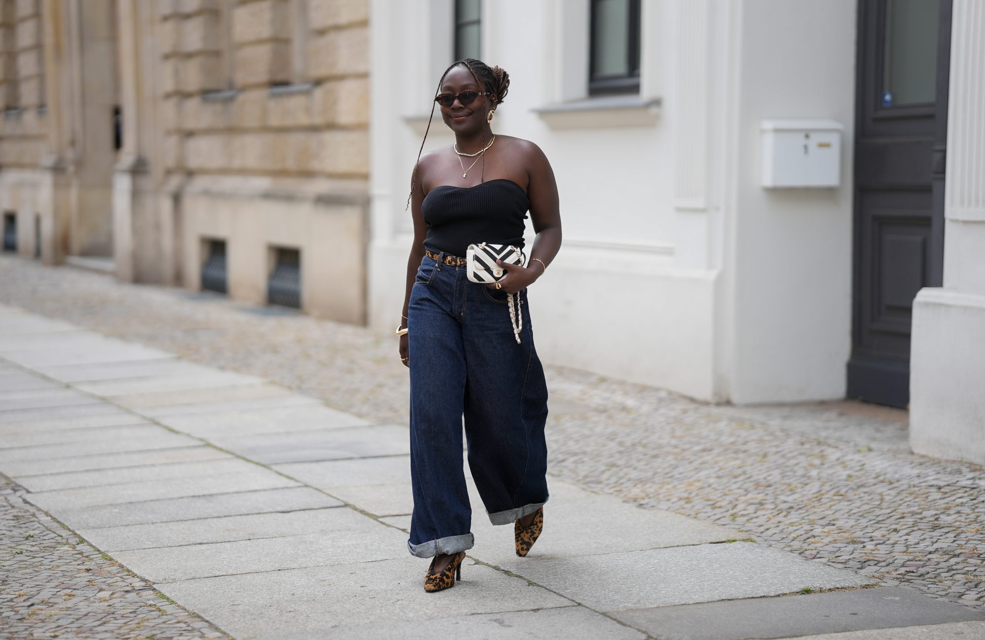 BERLIN, GERMANY - JULY 4: Lois Opoku is seen wearing a black ribbed strapless top with a sweetheart neckline from Uniqlo; dark blue high waist wide leg denim jeans with turned up cuffs from Levi’s; a slim leopard print belt from H&M Studio; leopard print pointed toe Mary Jane pumps from H&M Studio; a Chanel Mini CC bicolor chevron stitched lambskin flap shoulder bag with a gold tone chain strap; a gold collar necklace from Alexis Bittar and a gold pendant necklace from Bruna the Label; gold hoop earrings from ASOS; gold cuff bracelets and a gold chain bracelet from Petite Villa Mauresque; rings from Pandora and Maria Black; oval tortoiseshell frame sunglasses with dark tinted lenses from Corlin during the Berlin Fashion Week Spring/Summer 2026 on July 4, 2026 in Berlin, Germany. (Photo by Moritz Scholz/Getty Images)