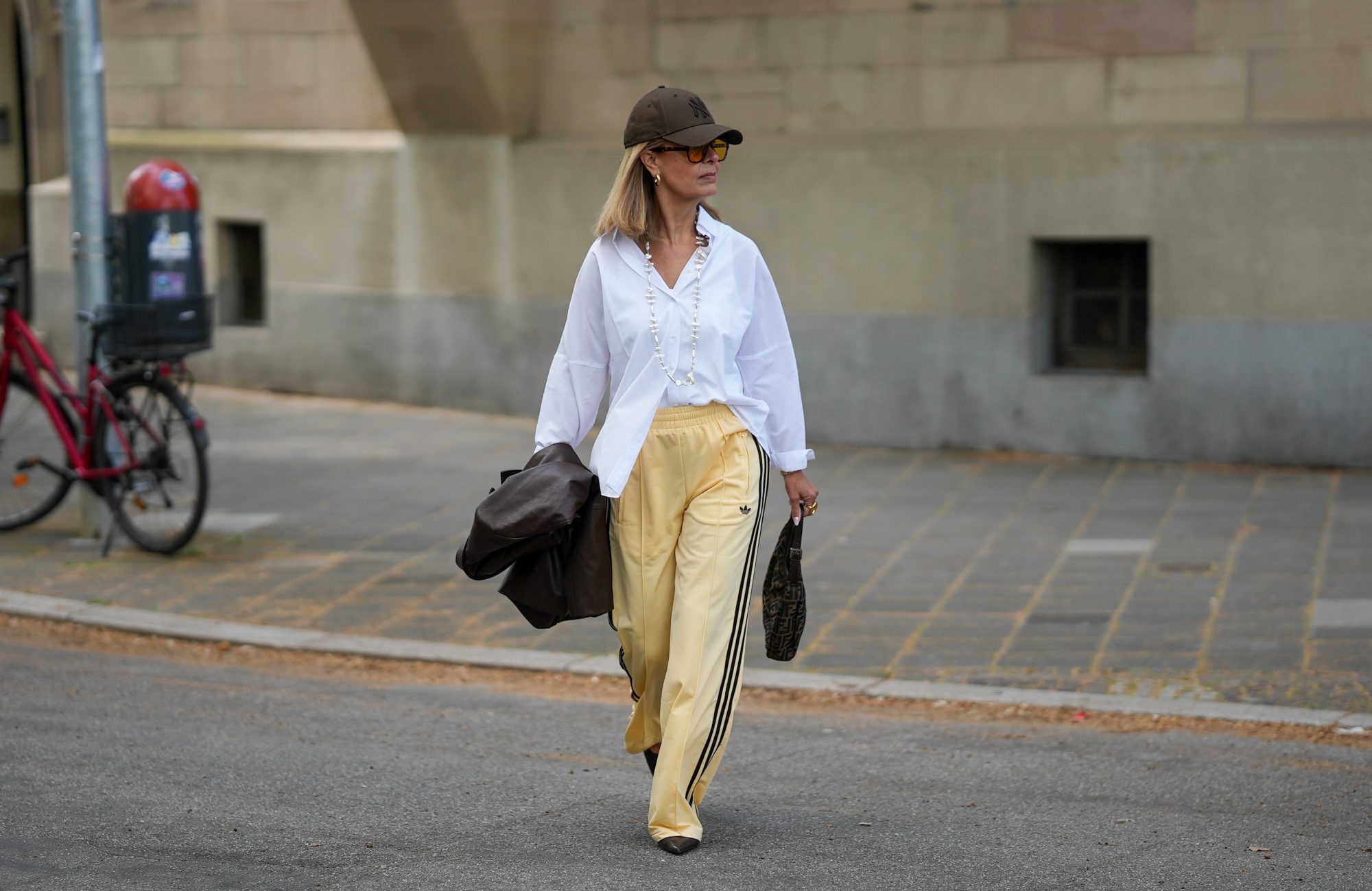 FRANKFURT AM MAIN, GERMANY - APRIL 21: Yvonne Schuppler-Kammerer is seen holding a dark brown leather blazer from Stradivarius; wearing an oversized white button down shirt from Zara; pale yellow wide leg track trousers with black three-stripe detail from Adidas; black pointed toe slingback pumps from H&M; a dark brown FF monogram canvas "Fendi Zucchino Half Moon Baguette Bag" from Fendi; a "Grace" gold wristwatch from Kapten & Son; a long pearl necklace from Zara; gold chunky hoop earrings and multiple gold rings from H&M and Scandivv; a nude manicure; oversized amber tinted square frame sunglasses from Bershka; a dark brown New York Yankees baseball cap from New Era; her blonde straight hair is worn loose; with natural make-up on April 21, 2026 in Frankfurt am Main, Germany. (Photo by Moritz Scholz/Getty Images)