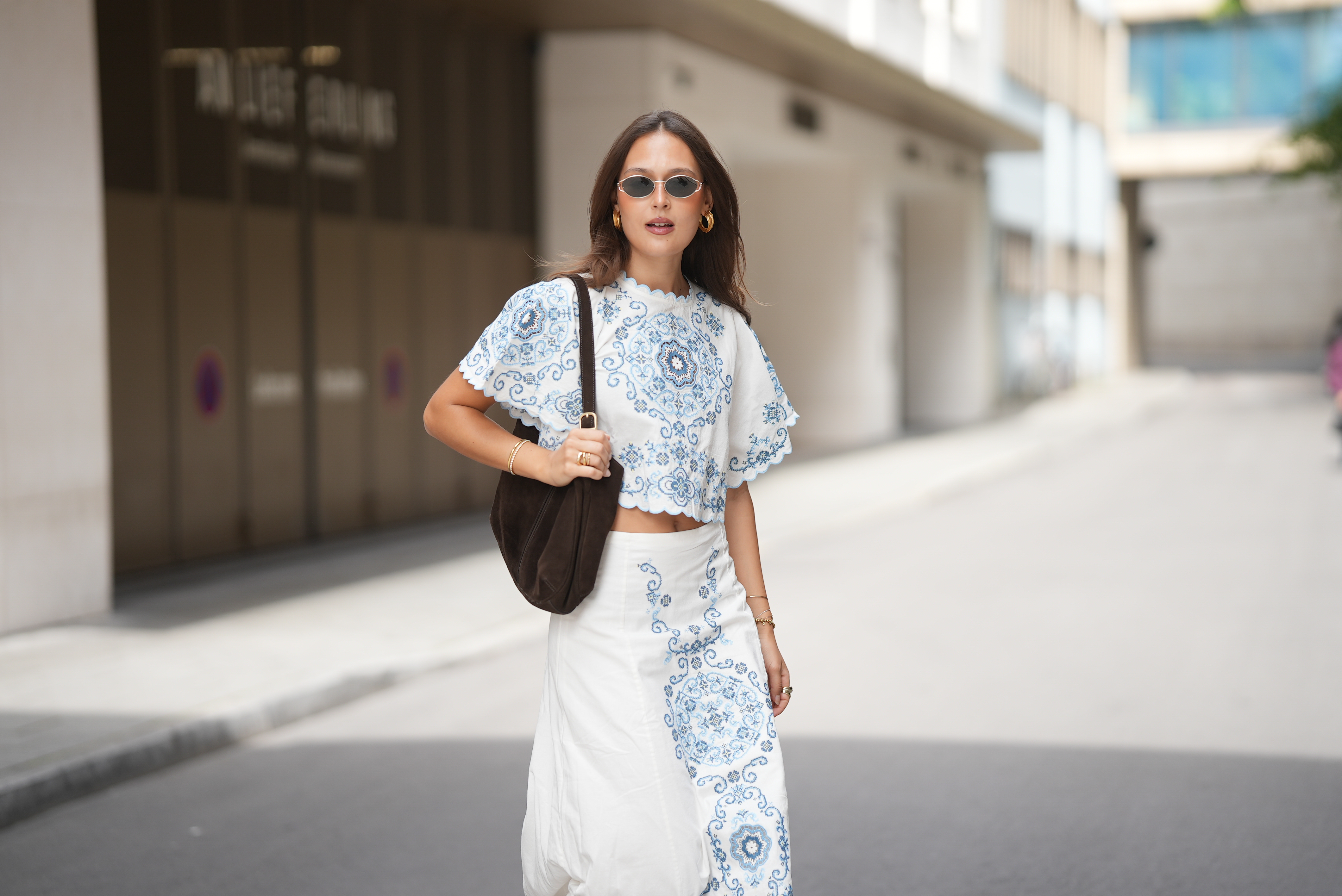 MUNICH, GERMANY - JUNE 16: Amanda M&uuml;hle is seen wearing a white cropped blouse with blue embroidered motifs, wide short sleeves and scalloped edges from S&eacute;zane; a white high waist midi skirt with blue embroidered motifs from S&eacute;zane; a dark brown suede shoulder bag with an adjustable strap and gold tone hardware from Rouje; gold hoop earrings, gold bracelets and gold rings; oval metal frame sunglasses with dark tinted lenses from Jimmy Fairly on June 16, 2026 in Munich, Germany. (Photo by Moritz Scholz/Getty Images)