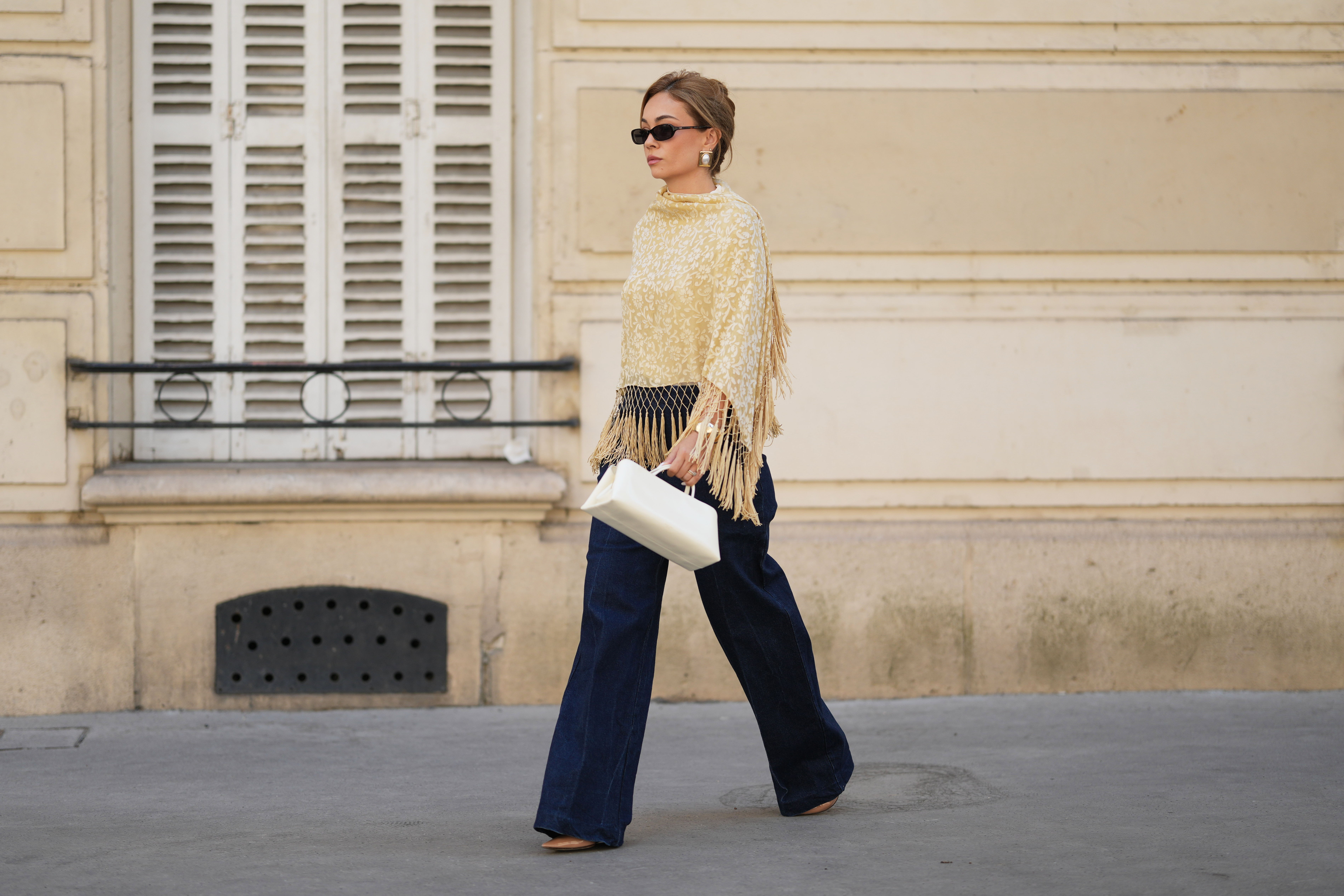 PARIS, FRANCE - AUGUST 17: Merve Gorgoz wears brown hair tied back with hair part, sunglasses, a beige yellow and white floral patterned vintage shawl with long knotted fringe, dark blue high-waisted wide-leg jeans denim pants by Sezane, a structured white handbag by Verafied, brown pointed-toe heels shoes by Jimmy Choo, during a street style fashion photo session, on August 17, 2026 in Paris, France. (Photo by Edward Berthelot/Getty Images)