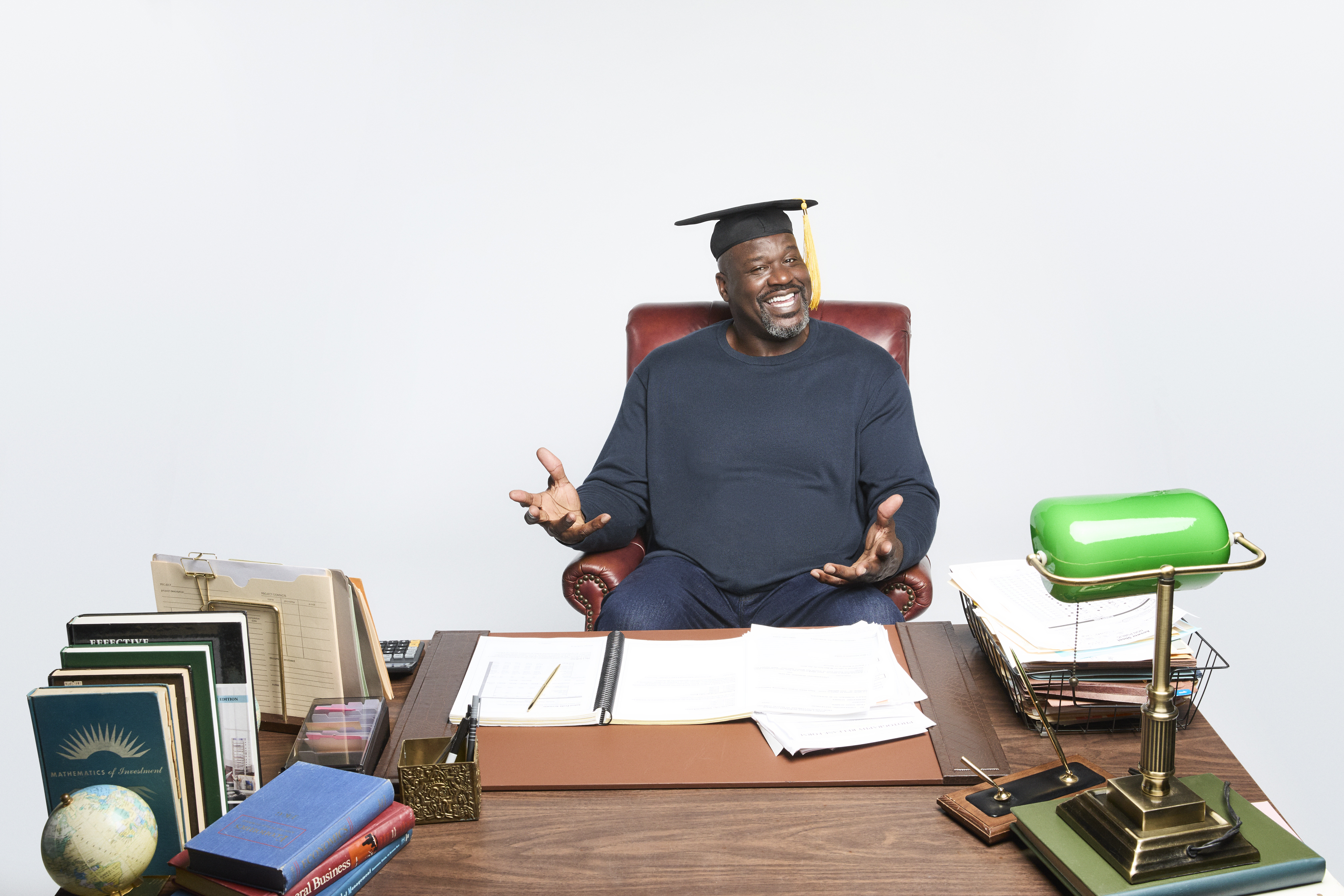 Shaquille O'Neal in cap and gown behind a school desk