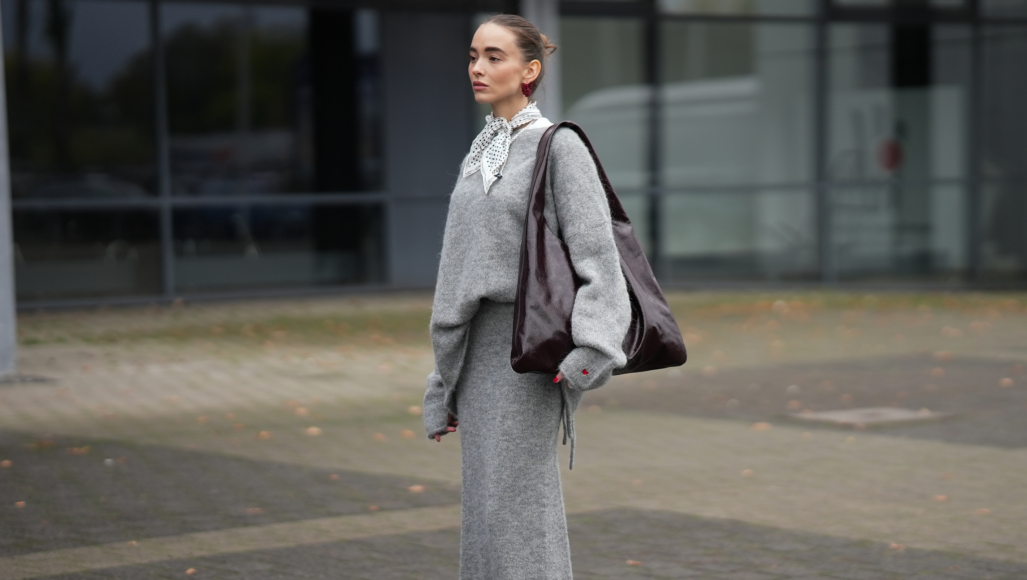 DUESSELDORF, GERMANY - OCTOBER 14: Evi Wave is seen wearing an oversized boatneck grey pullover by Aylin Koenig x Anita Hass with a slouchy fit, dropped shoulders and elongated sleeves made of a soft alpaca mix, it features an embroidered red heart one cuff; a full-length tube grey skirt by Aylin Koenig x Anita Hass in matching soft alpaca mix, with straight silhouette with a slightly flared hemline forming a co-ord set with the top; under the top she layered a white crewneck t-shirt; she carries an oversized shoulder hobo bag by Arket in high-shine patent leather in burgundy with a slouchy body; a pair of pointed-toe beige slingback kitten heels in monogram canvas by Gucci, featuring a brown logo-jacquard pattern, the GG motif with a low stiletto heel and brown leather piping around the edges, these worn with white ankle socks; on her fingers she wears silver rings from Ariane Ernst and Fossil; her nails are polished in red; she wears a silk carr&eacute; Fraas scarf with a polka-dot print in monochrome black and white tones, tied in a classic French knot at the collar; she wears a pair of sculptural beaded earrings handmade with red glass beads by Kroon02; her brown hair is styled in a sleek, low bun on October 14, 2025 in Duesseldorf, Germany. (Photo by Moritz Scholz/Getty Images)