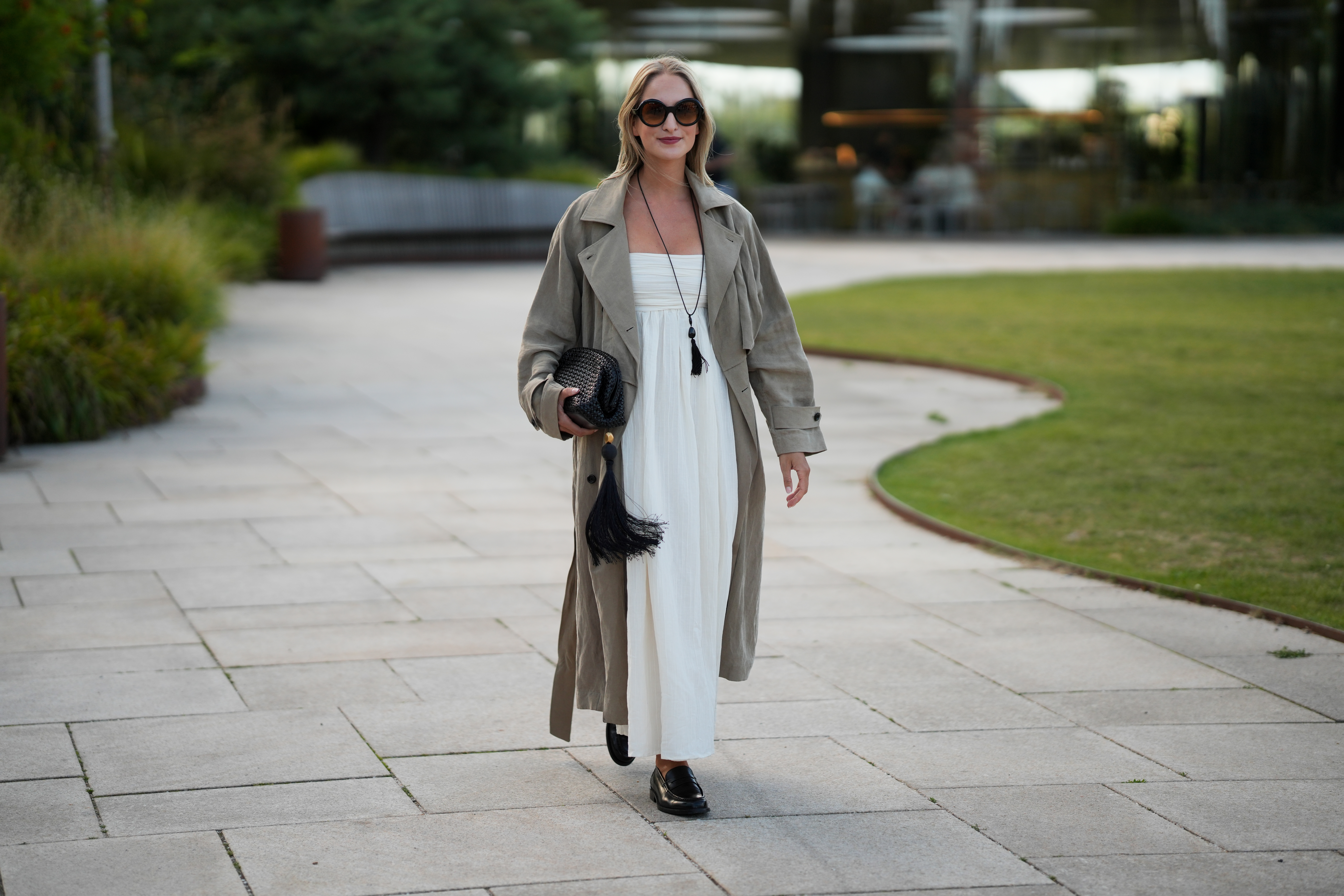 Louisa Theresa Grass wears oversized round sunglasses, a long taupe trench coat over a white strapless maxi dress, a long black tassel necklace, carries a black woven clutch with an oversized tassel and wears black loafers outside the Collina Strada show during day 4 of Copenhagen Fashion Week Spring Summer 2027 on August 6, 2026 in Copenhagen, Denmark.