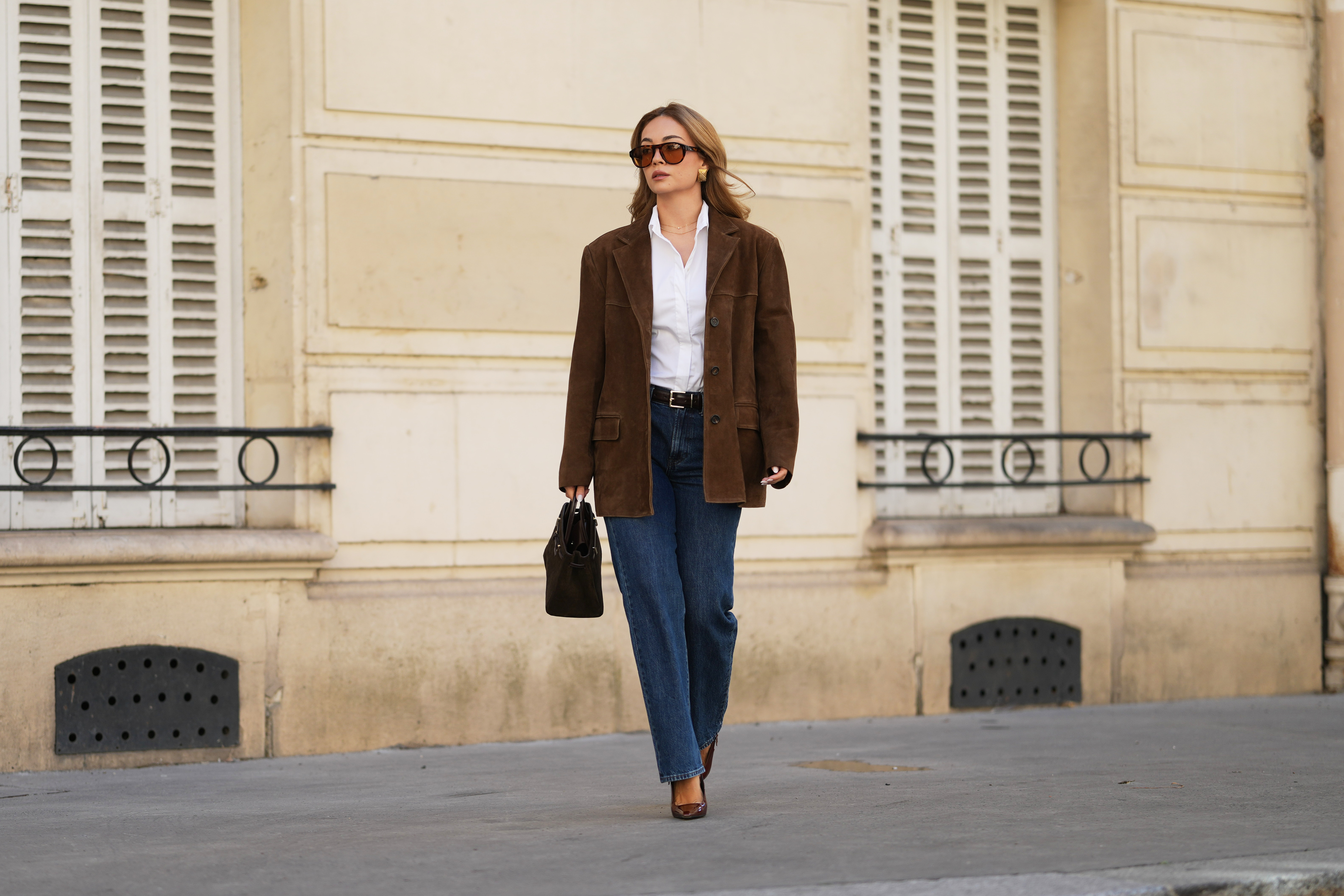 Merve Gorgoz wears brown long hair with center part, amber colored sunglasses with orange tinted lenses by Jimmy Fairly, golden square earrings, a brown suede oversize blazer jacket by Source Unknown, a white shirt, a leather belt, blue denim pants jeans by Mango, a dark brown khaki bag in suede by Demellier, brown pointed shiny patent leather shoes with high heels by YSL Saint Laurent, during a street style fashion photo session, on September 01, 2026 in Paris, France.